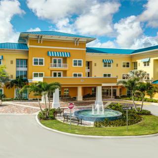 A bright yellow residential building with a fountain in front, surrounded by trees and a clear blue sky.
