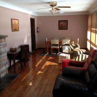 A cozy living room with hardwood flooring, featuring a sofa, chairs, a fireplace, and a ceiling fan. Sunlight streams through the windows.