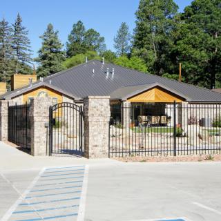 A fenced building with stone columns and a gated entrance in front of a parking lot, surrounded by trees.