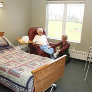 Two elderly people sitting in a nursing home room, one in a recliner and the other appearing to provide care.
