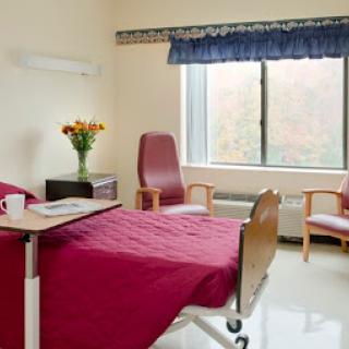 A hospital patient room featuring a bed, chairs, and a window with floral arrangements.
