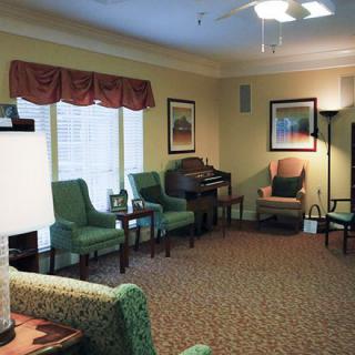 A cozy living room area with green chairs and a sofa arranged around a carpeted floor, featuring ambient lighting and windows with curtains.