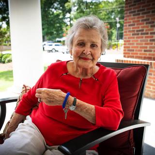An elderly woman in a red shirt is sitting on a porch, smiling at the camera.