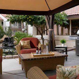 A cozy outdoor patio with wicker furniture and cushions under a gazebo, surrounded by a garden setting.