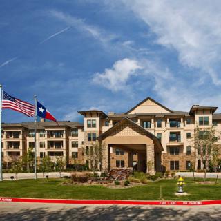 A modern building with American and Texas flags displayed at the entrance, surrounded by well-maintained landscaping under a clear blue sky.