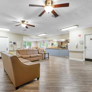 A spacious living room with wooden flooring, featuring sofas and ceiling fans, illuminated by natural light through large windows.
