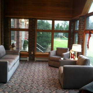 A cozy living room with a sofa, chairs, and a lamp, overlooking a green lawn through large windows.