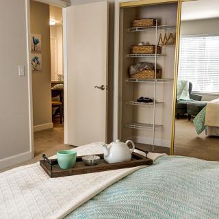 A cozy bedroom featuring a neatly arranged tray with a teapot, open closet showcasing shelves, and a glimpse of an adjoining room.
