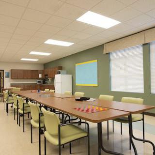 A spacious meeting room with a large conference table and chairs, featuring a kitchen area in the background.