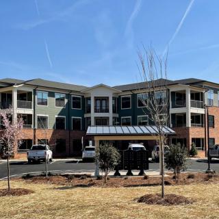 A modern residential apartment building with multiple floors and a landscaped area in front, set against a clear blue sky.