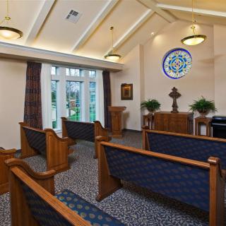A small chapel interior with wooden pews, stained glass windows, and warm lighting.