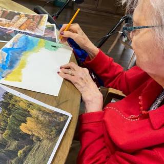 An elderly woman engaged in watercolor painting indoors, drawing inspiration from landscape photographs.
