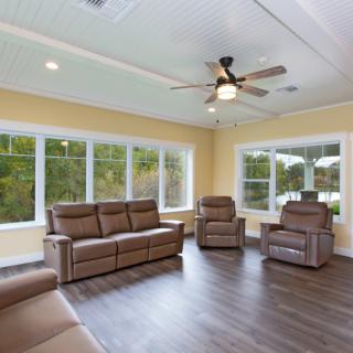 A spacious living room with large windows, brown leather furniture, and a ceiling fan.
