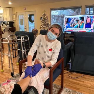 A caregiver wearing a mask and gloves tends to a patient in a nursing home setting, with a television on in the background.