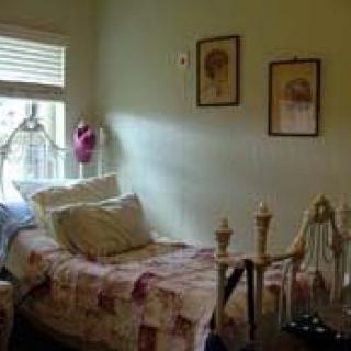 A cozy vintage-style bedroom with a single bed, pillows, framed art on the wall, and soft lighting from a window.