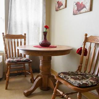 A cozy dining nook featuring a wooden table with two matching chairs, adorned with cushions and a red vase holding a single flower.