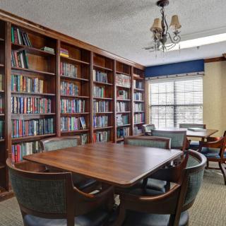 A cozy library room with bookshelves, a central wooden table surrounded by chairs, and natural light coming through the window.