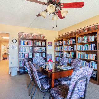 A cozy reading room with bookshelves lined with a variety of books, a table surrounded by cushioned chairs, and a ceiling fan overhead.