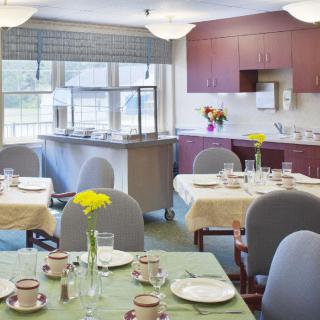 A cozy dining room with neatly set tables, complete with floral decorations and a kitchen area in the background.