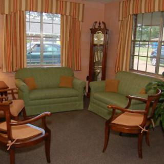 A cozy living room with green sofas, wooden chairs, and a grandfather clock by the windows.