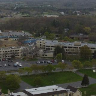 An aerial view of an urban area featuring multiple buildings, a parking lot, and patches of green space under an overcast sky.