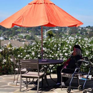 A serene outdoor patio scene with an orange umbrella, flowers, and a person sitting next to a wheelchair.