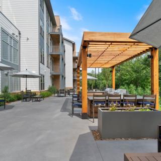 A modern residential outdoor patio with a wooden pergola, featuring tables and chairs under a clear blue sky.