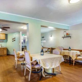 A cozy dining area with neatly arranged tables and chairs, decorated with tablecloths and decorative bottles, featuring a warm wooden floor.