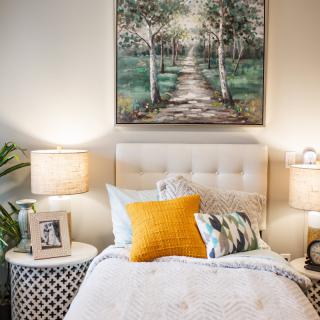 A cozy bedroom with a neatly made bed, featuring colorful pillows and an elegant landscape painting above the headboard. Warm lighting from bedside lamps and a potted plant add to the inviting atmosphere.