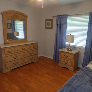 A cozy bedroom featuring a wooden dresser with a mirror, a bedside table with a lamp, and blue curtains framing the window.
