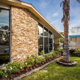 A modern building with a stone facade and large glass windows, surrounded by manicured landscaping featuring a palm tree.