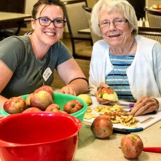 A young woman and an elderly woman are smiling while preparing apples together in a kitchen.