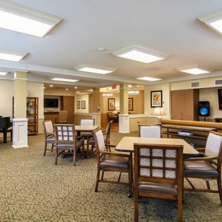 A spacious community lounge with tables and chairs, featuring warm lighting and carpeted flooring.