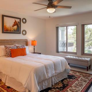 A cozy bedroom with a neatly made bed, decorative pillows, and a large rug, featuring natural light from windows and tasteful artwork on the walls.