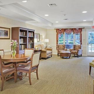 A cozy living area with bookshelves, a table, chairs, and sofas, featuring large windows and warm decor.