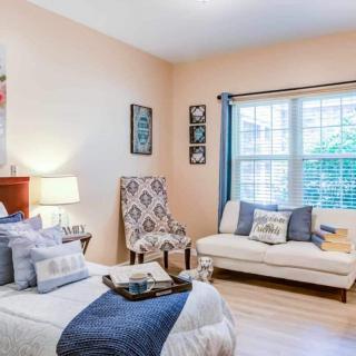 A cozy bedroom with a bed, armchair, and sofa, featuring light colors and elegant decor.