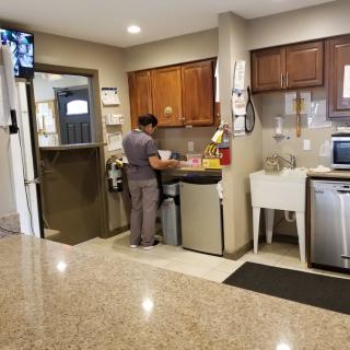 A person cleaning in a kitchen area of a facility, with cabinets, a sink, and a microwave visible.
