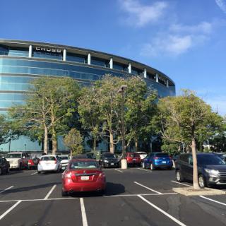A modern building with a curved glass facade surrounded by trees and parked cars on a sunny day.