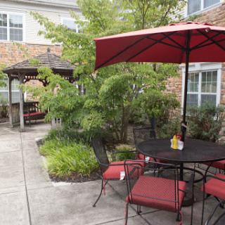 A cozy patio area with red-cushioned chairs, a matching umbrella, and a gazebo surrounded by greenery.