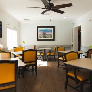 A bright and inviting dining room with multiple tables and chairs, featuring a ceiling fan and a landscape painting on the wall.