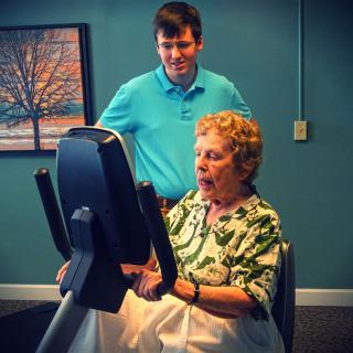 An elderly woman is assisted by a man while using fitness equipment for exercise.