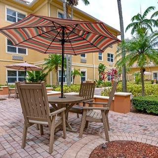 A cozy outdoor patio area with table and chairs under a striped umbrella, surrounded by lush greenery and palm trees near a residential building.