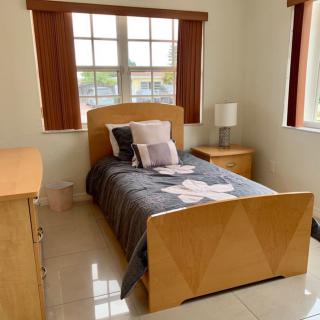 A well-lit bedroom featuring a single bed with a dark comforter, wooden furniture, and large windows.