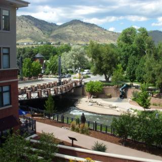 A scenic view of a river flowing under a bridge with mountains in the background, surrounded by trees and buildings.