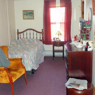A cozy bedroom featuring a bed, an upholstered chair, and a dresser with a mirror, complemented by a window with red curtains.
