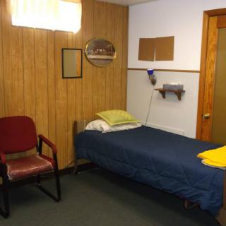 A simple bedroom with wood paneling, featuring a single bed and a chair.