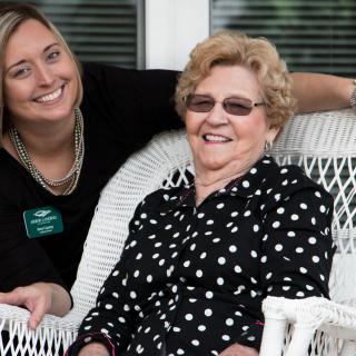 A smiling elderly woman in a polka dot shirt sits on a white wicker chair next to a younger woman outdoors.