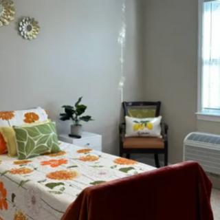 A cozy bedroom with a floral-themed single bed, featuring decorative pillows and a small chair by the window.