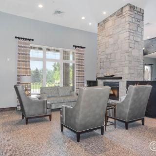 A modern living room with a central fireplace surrounded by armchairs and a sofa, featuring large windows that let in natural light.