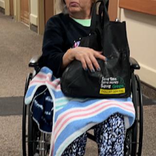 A patient sitting in a wheelchair, wrapped in a blanket and holding a handbag.